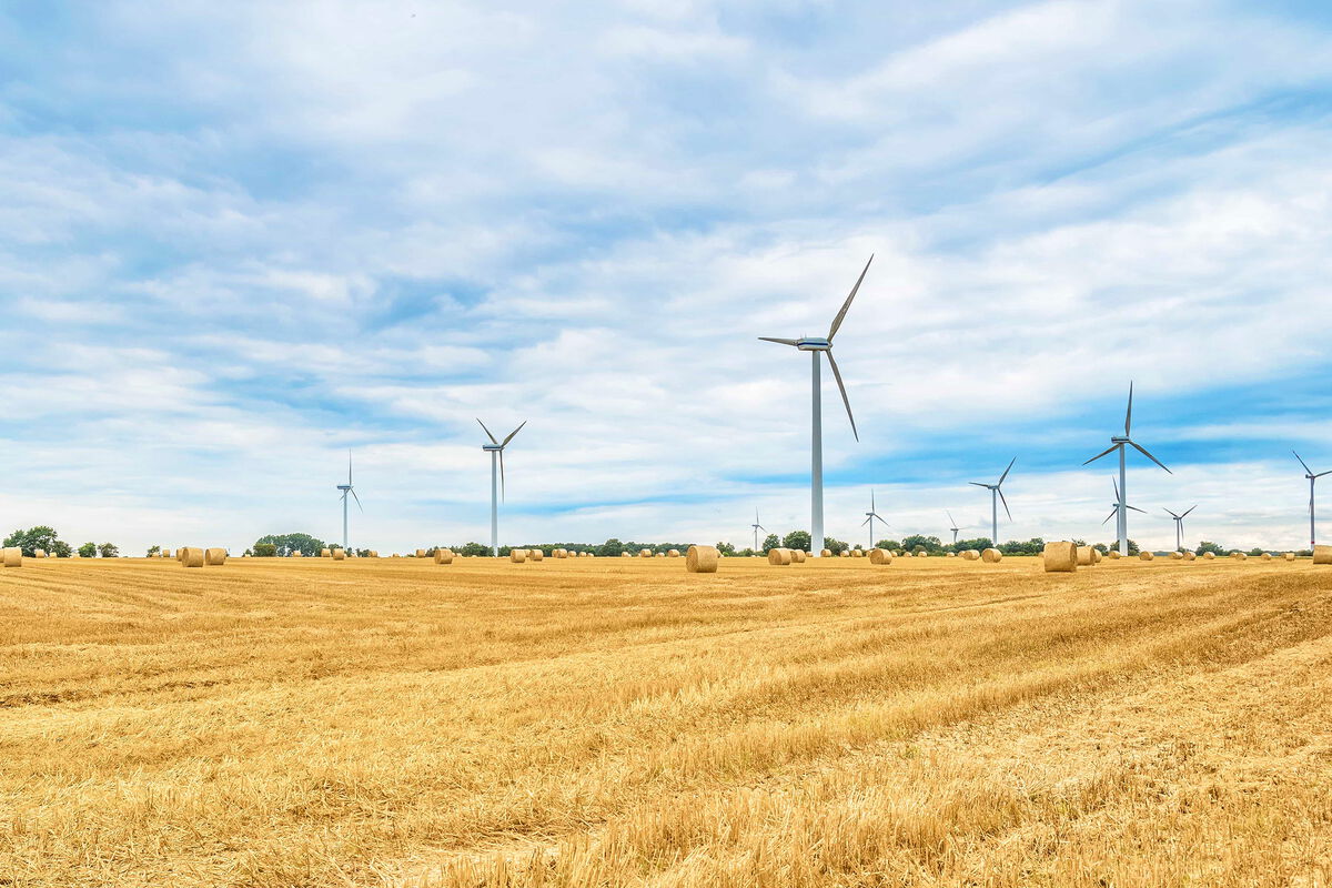 abgemähtes Getreidefeld, im Hintergrund mehrere Windräder sowie Strohballen, blauer Himmel mit Wolken